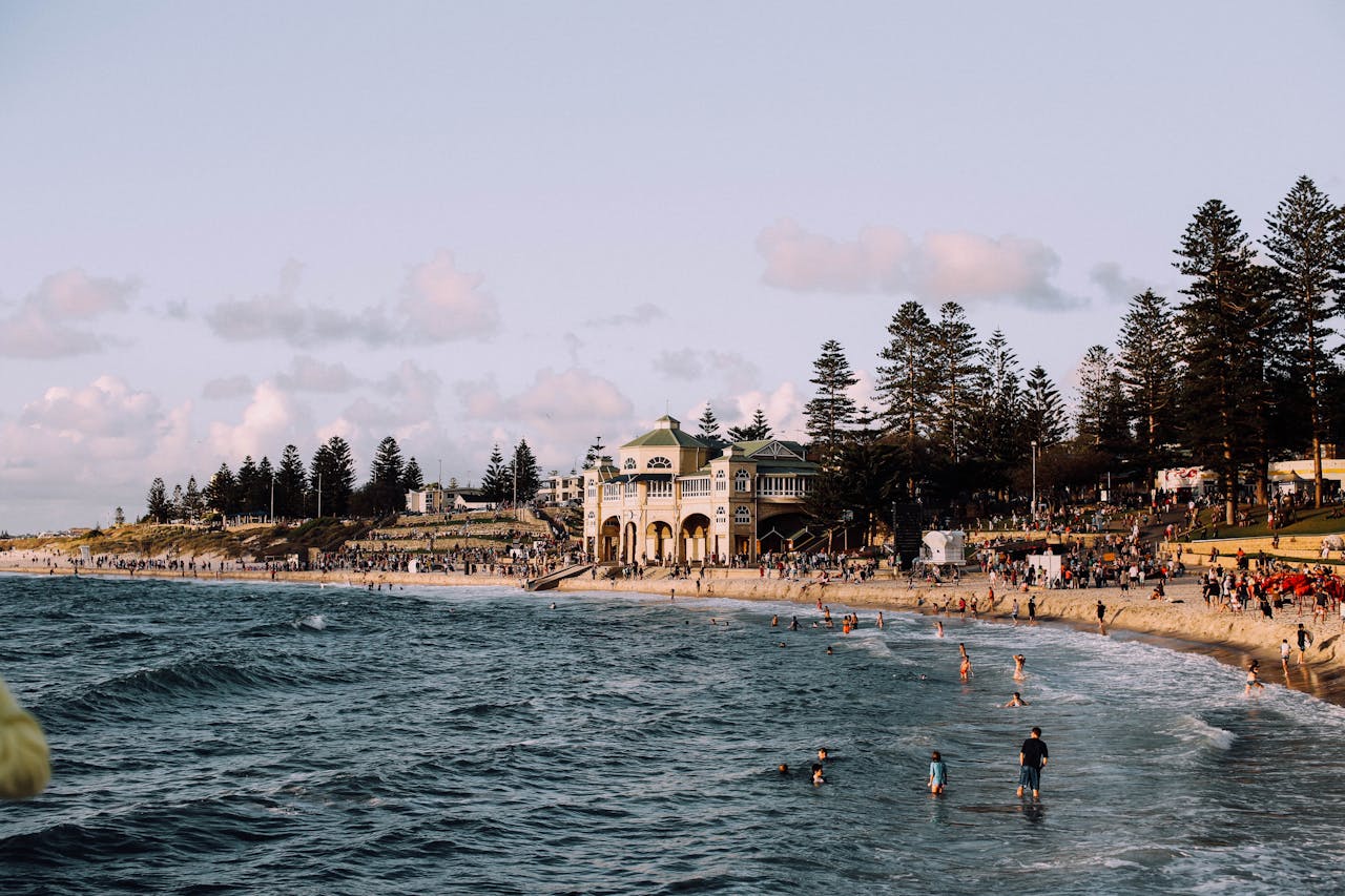 Anonymous tourists walking and swimming in sea with sandy shore with coniferous trees under cloudy sky